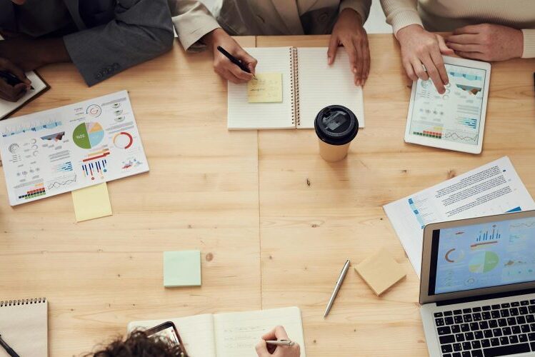 Team meeting around a table with charts, notebooks, a tablet, and a laptop displaying analytics dashboards