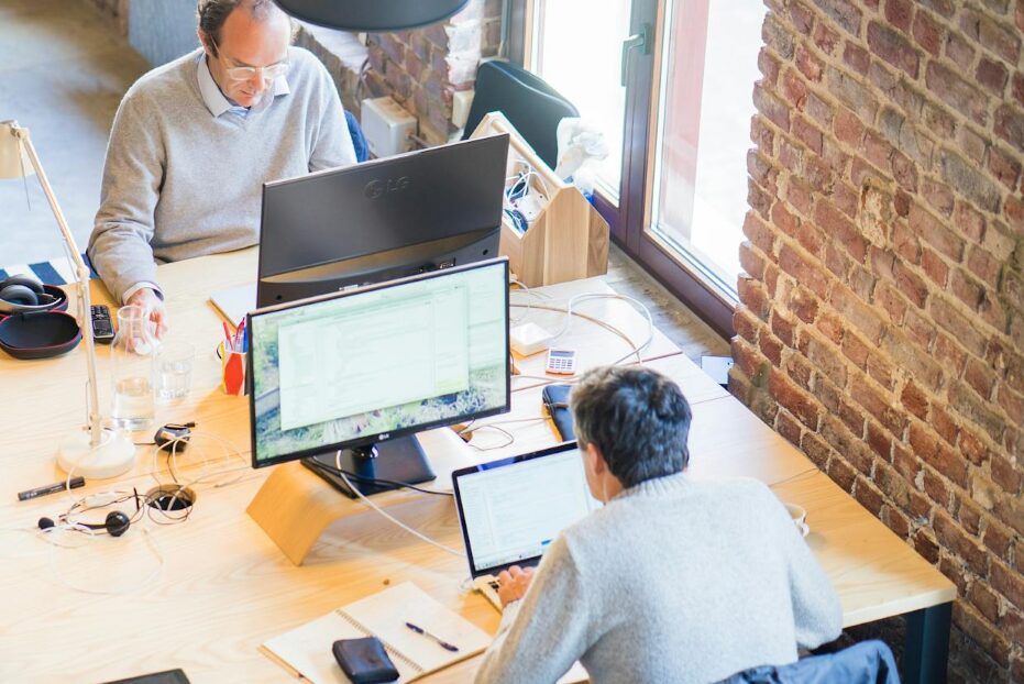 Two coworkers working at a shared desk with multiple computer monitors in an office
